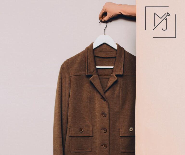 Hand houdt een bruine maatwerkjas op een witte kleerhanger tegen een zachte beige achtergrond met logo Interieurwerken Janssens